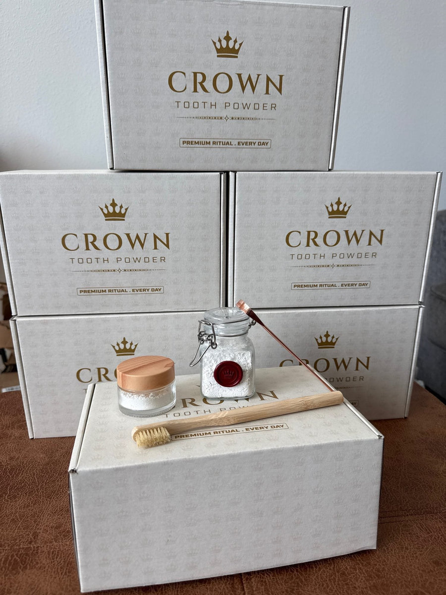 Stack of printed Crown Tooth Powder boxes with the glass jar, travel jar, bamboo brush, and copper tongue scraper arranged in front.