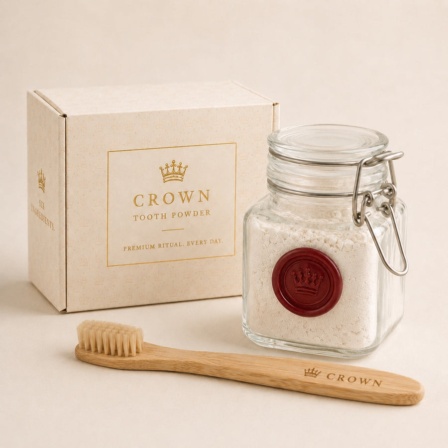 A Crown Tooth Powder box, a sealed glass jar of white powder with a burgundy wax crown seal, and a bamboo boar bristle toothbrush on a warm neutral background