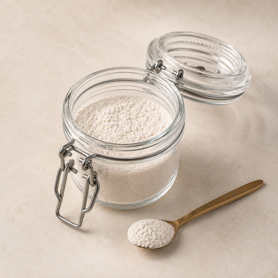 An open glass jar of tooth powder beside a brass spoon holding a small scoop of the powder on a warm neutral surface